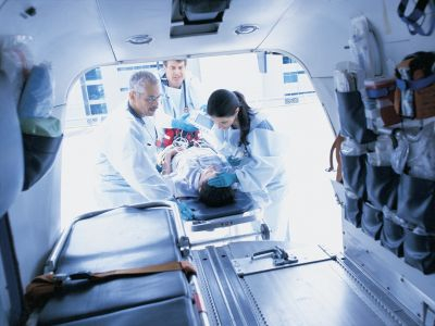 A group of healthcare workers in white coats is seen assisting a patient in an ambulance setting.